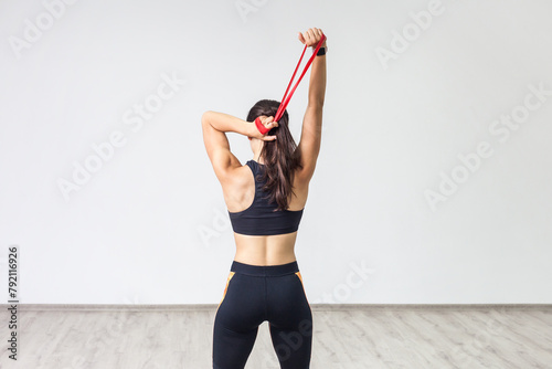 Back view of young sporty woman wearing black tank top and leggings, doing exercise for triceps with latex resistance band. Full length indoor shot against white wall. photo