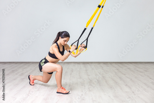 Portrait of strong woman wearing sportswear doing lunges having workout for legs and glutes using trx in gym, taking care of her physical shape. Indoor shot against white wall background photo