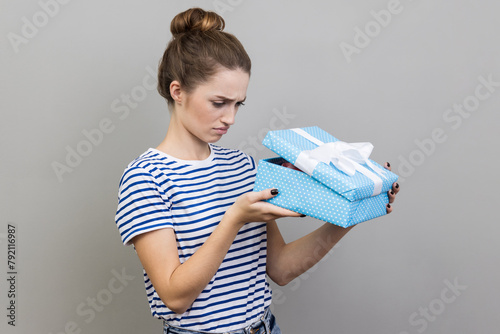 Portrait of sad woman wearing striped T-shirt unboxing blue gift box, looking inside with upset facial expression, bad birthday present. Indoor studio shot isolated on gray background.