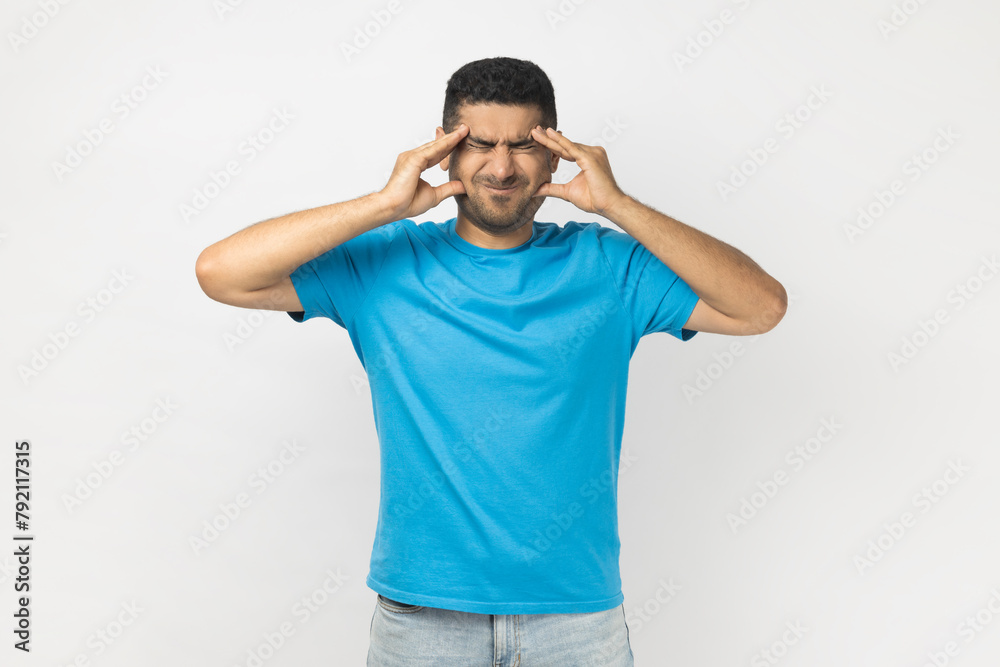 Portrait of sick unhealthy ill unshaven man wearing blue T- shirt standing massaging temples, suffering headache, feels fatigue. Indoor studio shot isolated on gray background. Portrait of sick unhealthy ill unshaven man wearing blue T- shirt standing massaging temples, suffering headache, feels fatigue. Indoor studio shot isolated on gray background.