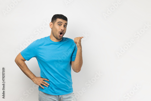 Portrait of amazed shocked young adult unshaven man wearing blue T- shirt standing indicating at advertisement area, copy space for promotion. Indoor studio shot isolated on gray background.