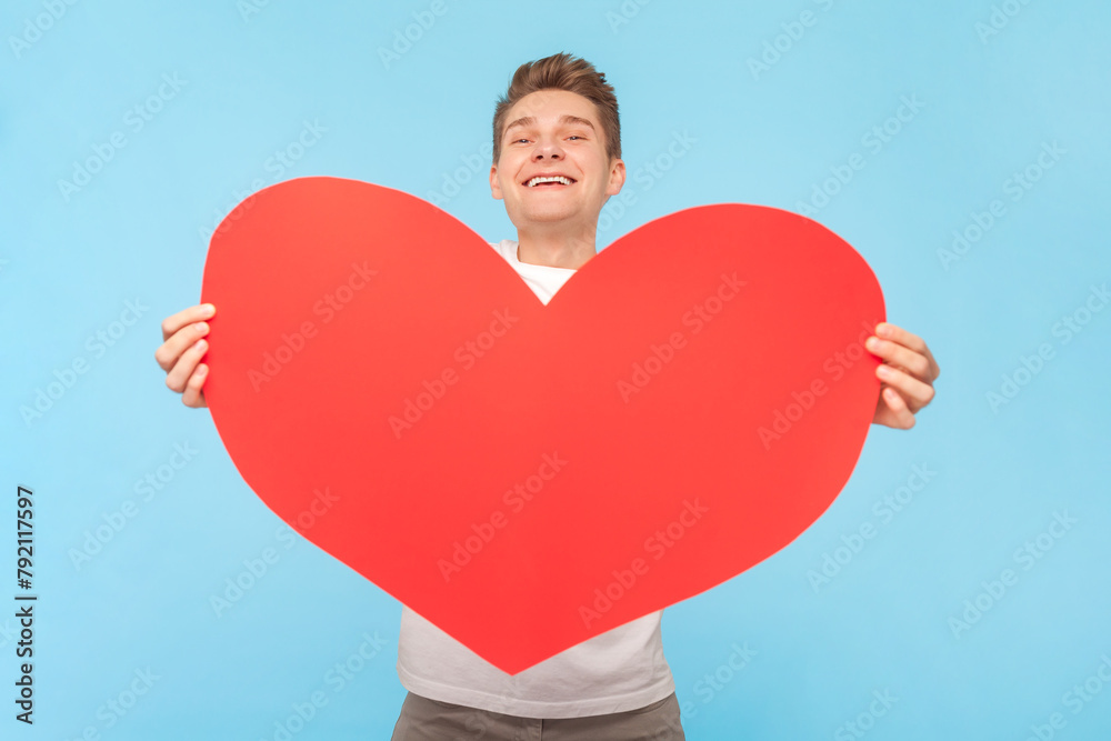 Portrait of satisfied smiling pretty man wearing white t-shirt holding big red heart, expressing love and devotion. Indoor studio shot isolated on blue background Portrait of satisfied smiling pretty man wearing white t-shirt holding big red heart, expressing love and devotion. Indoor studio shot isolated on blue background