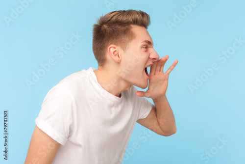 Side view portrait of angry aggressive man wearing white t-shirt standing screaming loud, making announce, arguing with somebody. Indoor studio shot isolated on blue background