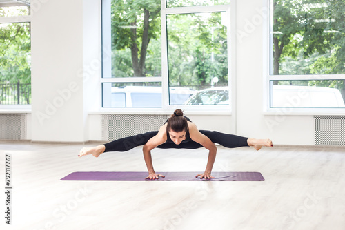 Full length portrait of strong athletic young woman doing yoga pose on hands, balance, healthy lifestyle, wearing black pants and top. Indoor shot in gym near big windows.