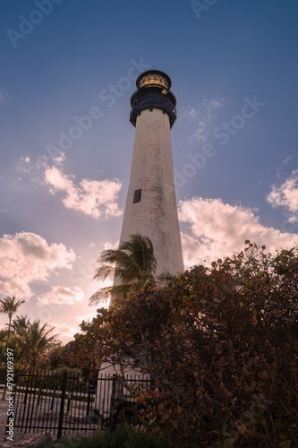 A white lighthouse against deep blue sky with white clouds tinted with orange pink sunset sun rays with lavish colorful green red orange violet plants trees bushes, palm trees in the foreground
