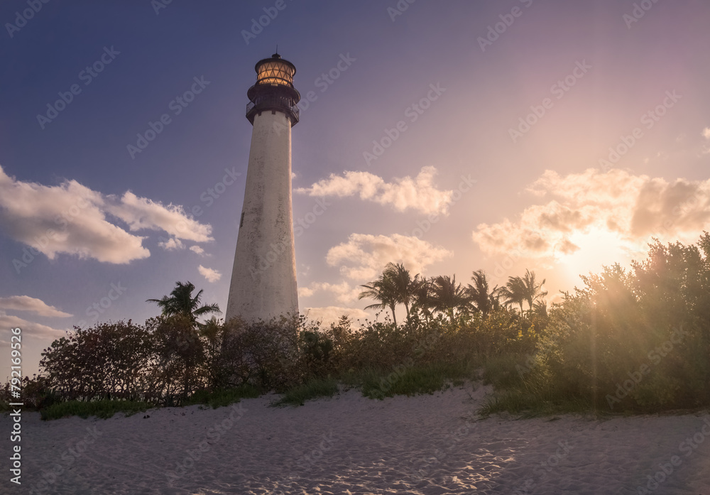 A white lighthouse against deep blue sky with setting sun tinting white clouds with orange pink light rays with white sand of a beach and green bushes and palm trees in the foreground