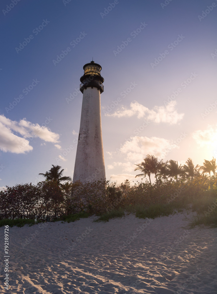 A white lighthouse against deep blue sky with white clouds tinted with orange pink sunset sun rays with lavish colorful green red orange violet plants trees bushes, palm trees in the foreground