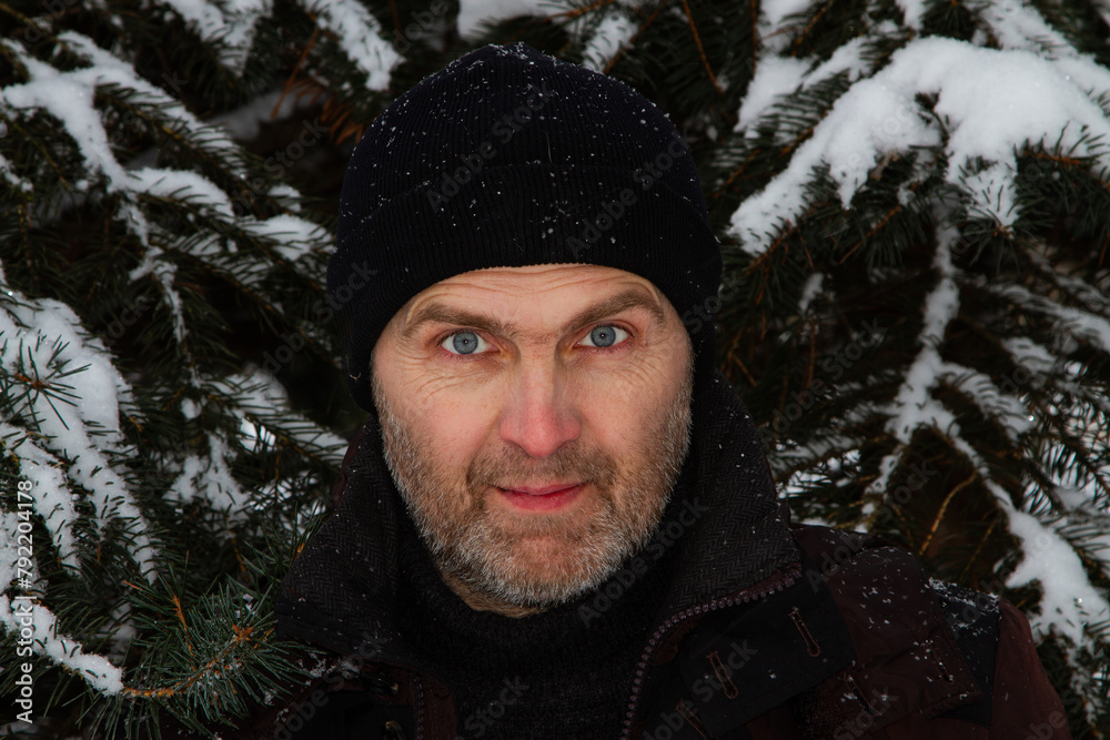 Man with beard wearing hat in nature. Close-up portrait of a handsome unshaven man after 40 years old wearing a black hat looking at the camera in a winter snow-covered forest.