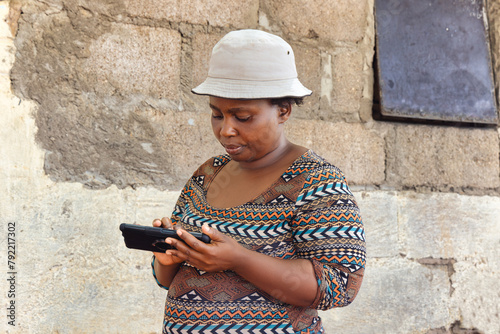 young african chubby woman with a hat holding her cellphone in the poor township, informal settlement