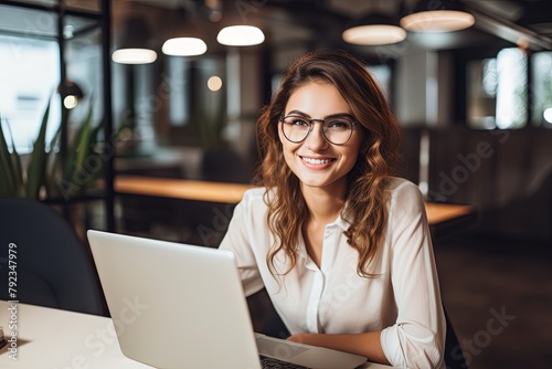 Photo of beautiful happy woman looking at camera while sitting at office. using laptop