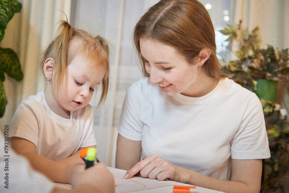 Young mother or babysitter, little daughter drawing at table in room. Painting, doing homework, family enjoying leisure at home Young mother or babysitter, little daughter drawing at table in room. Painting, doing homework, family enjoying leisure at home
