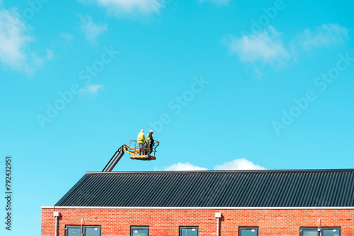 Roofers working off telescopic boom lift while inspecting and replacing roof tiules on a  multistorey residential building against blue sky with space for test. Working at height safely poster concept