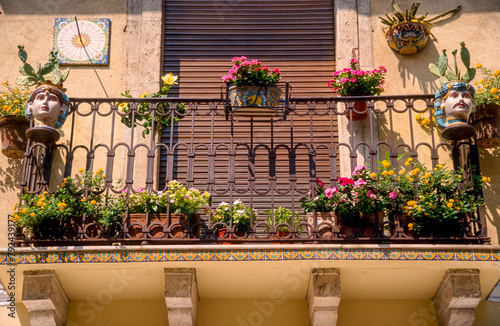 Ein mit bunten Zierblumen geschmückter kleiner Balkon mit schmiedeeiserner Balustrade in mediterranem Stil bei Sonnenschein und geschlossener Balkontür