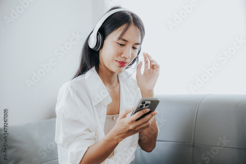 Young Asian woman listening to music on the sofa in the living room at home Happy Asian woman using mobile smartphone Wear a headset and sit on the sofa.