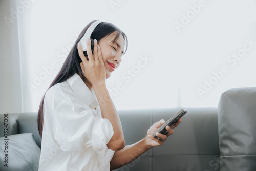 Young Asian woman listening to music on the sofa in the living room at home Happy Asian woman using mobile smartphone Wear a headset and sit on the sofa.