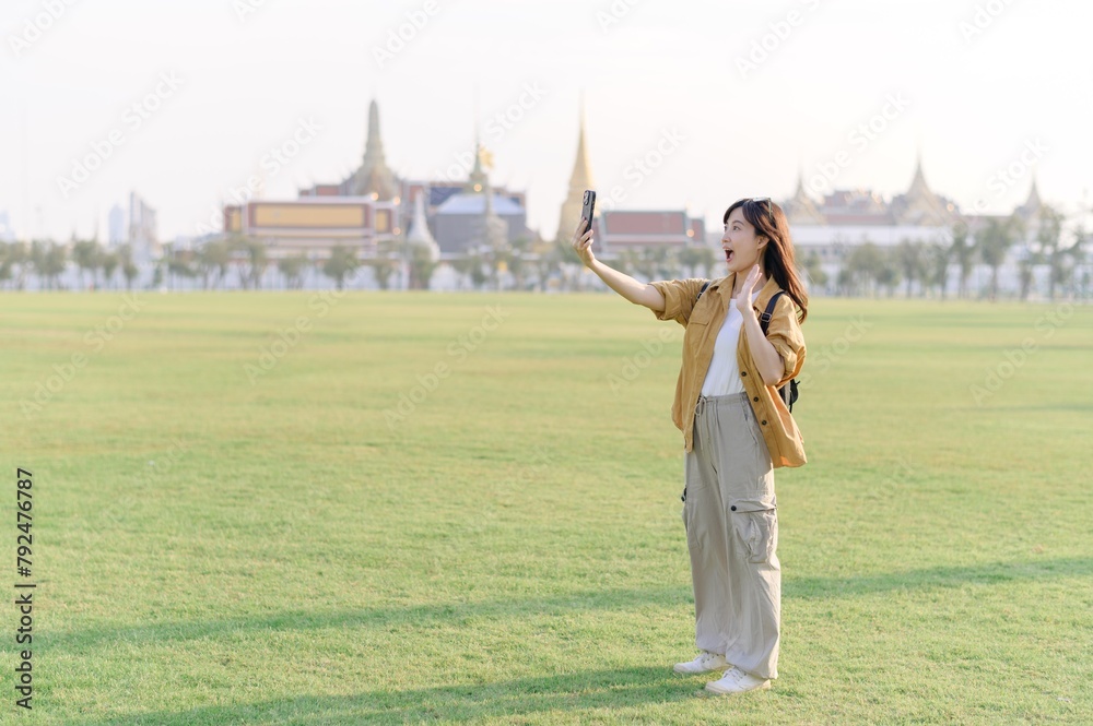 Fototapeta premium Traveler asian woman in her 30s take a livestream on smartphone while explores Wat Phra Kaew emerald Buddha. Share the wonders of Thai heritage through her journey.