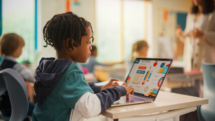 Teacher Explaining a Math Lesson to a Classroom Full of Bright Diverse Children in Primary School. Little Black Boy Browsing a Presentation on a Laptop Computer