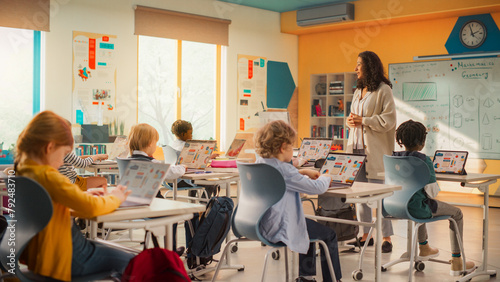 Group of Tech-Savvy Primary School Children Working on Their Laptop and Tablet Computers in Class. Young Female Teacher Educating Kids on Internet Safety, Basic Programming Languages and Formulas