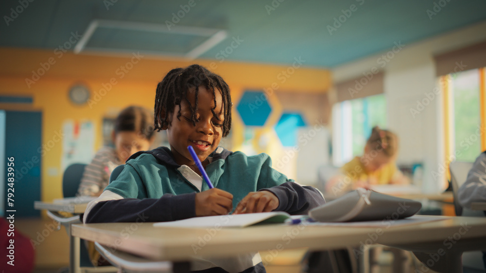 Portrait of a Cute Little African Boy with Stylish Hair Sitting Behind a Desk in Class in Elementary School. Young Pupil is Focused on a Lecture, Listening to a Teacher with Other Kids