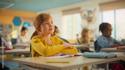 Portrait of a Cute Smiling Girl with Bright Ginger Hair Sitting Behind a Desk in Class in Elementary School. Young Pupil is Focused on a Lecture, Listening to a Teacher with Other Kids photo