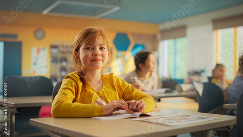 Portrait of a Cute Smiling Redhead Girl Sitting Behind a Desk in Class in Elementary School. Young Pupil is Looking at Camera, Smiling. Kids Educated in Modern Primary School photo