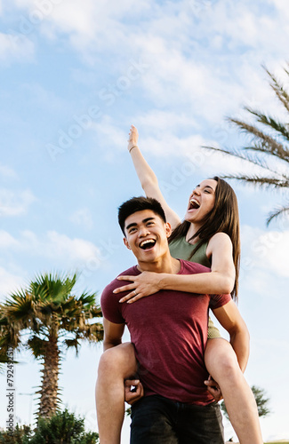 Vertical portrait of happy young multiracial couple having fun together on summer vacation. Joyful asian man piggybacking pretty woman outdoors. Love, happiness and friendship concept