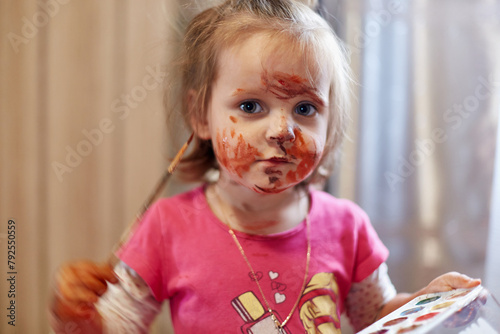 Charming child drawing and stains everything with paints holding color palette and paintbrush looking at camera with dirty colorful face