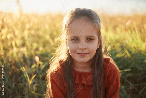 Charming brown haired Caucasian little girl wearing red shirt playing outdoor posing in sunny meadow adorable cute kid looking at camera with gently smile