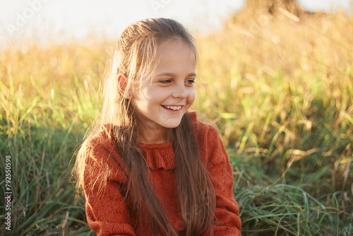 Laughing dark haired Caucasian joyful little girl sitting on grass in field looking away with happy smile enjoying outdoor walking on hot sunny summer day