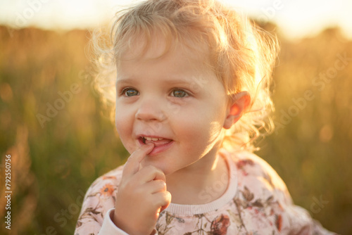 Adorable baby girl with blond wavy hair at sunny summer day looking away with curious happy face keeps mouth open holding finger on lips posing at sunset outdoor