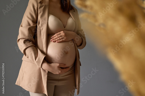 Unknown young pregnant girl in beige jacket and lingerie standing against gray wall background with dried flower stroking her belly with tenderness and love