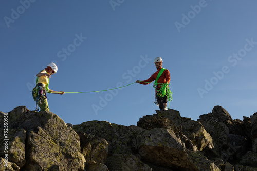 Two people are on a mountain, one of them is holding a rope. The other person is wearing a green harness