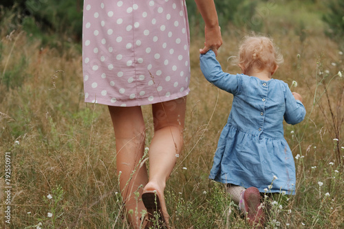 Unrecognizable woman legs walking in field on grass holding hand of infant little daughter wearing cute blue dress family walking in meadow on summer day