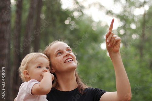 Cute little girl in mother hands looking away while mommy pointing at something interesting in forest young family spending time outdoor on summer day