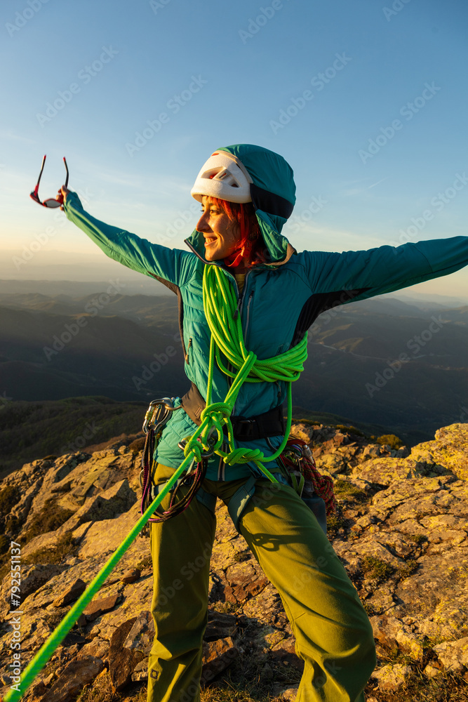 A woman in a green jacket is standing on a mountain with a green rope around her waist. She is smiling and she is enjoying the view - obrazy, fototapety, plakaty 