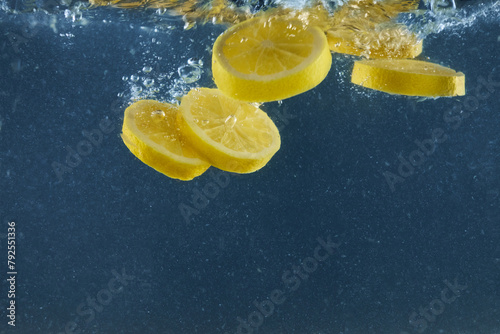 Yellow lemon slices falling in water for making cocktail isolated on blue background, empty space for advertisement content