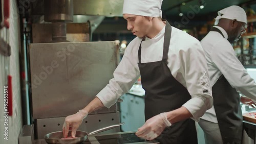 Male chef in apron, hat and gloves placing meat steaks on frying pan while preparing order in restaurant kitchenMale chef in apron, hat and gloves placing meat steaks on frying pan while preparing ord photo