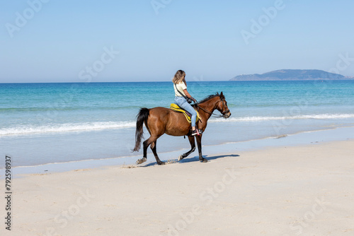 A female equestrian rides a brown horse along the shoreline with clear blue waters and a mountainous backdrop, capturing a moment of peaceful coexistence with nature