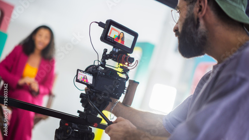 Film Set With A Young Black Female Host In A Pink Suit Engaging In A Lively Discussion, Captured By A Male Cinematographer Focusing Intently On His Camera Equipment, Surrounded By Colorful Backdrops. © Gorodenkoff