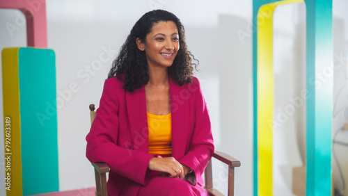 Young Black Female Host In Vibrant Pink Suit And Yellow Top, Smiling Engagingly On Colorful Set, Exemplifies Modern Videography And Dynamic Storytelling In Media Production.