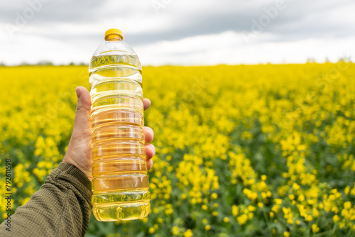 A bottle of rapeseed oil in a hand against the background of a yellow blooming rapeseed field. A bottle of rapeseed oil and blooming rapeseed fields.