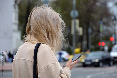 Girl with blond hair using smartphone on city street on blurred cars backgrounds