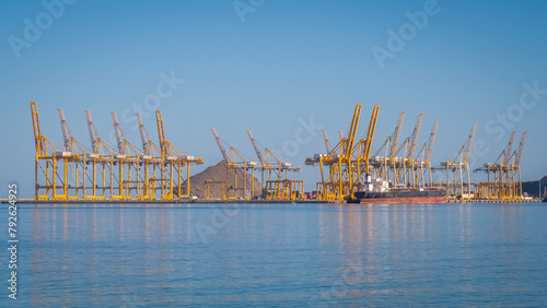 Port cranes loading container ship in  the Port of Fujairah, United Arab Emirates. Rows of yellow port cranes against blue sea and sky.