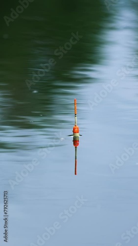 White and orange fishing bobber floating in the river. Unmoving float on still water on cloudy day. Vertical video