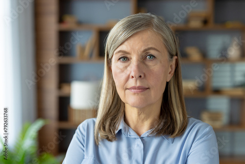 A professional portrait of a senior Caucasian businesswoman in a blue shirt, looking directly at the camera with a confident and calm expression in a well-lit office environment.