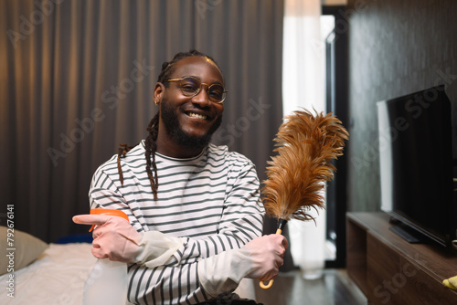Happy smiling young African guy holding a feather duster ready for cleaning his apartment