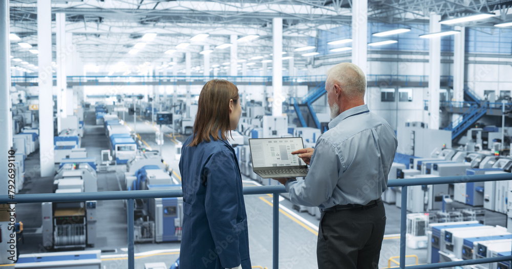 Obraz premium Two Multiethnic Engineers Standing on a Platform, Using Laptop Computer at an Electronics Factory. Machines are Undergoing Maintenance, Specialists Monitoring the Progress Through Online Software