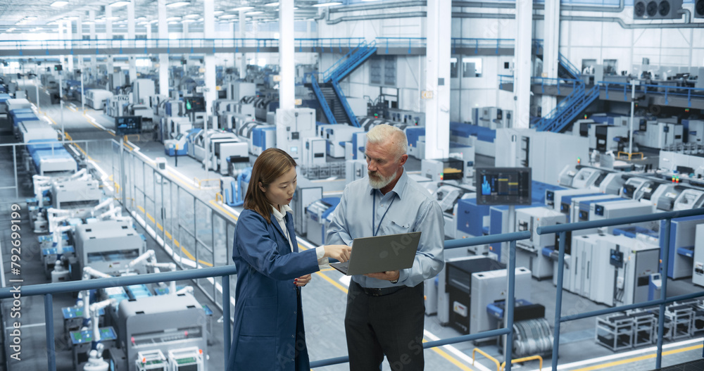 Experienced Male and Female Engineers Standing on a Platform, Using Laptop Computer and Discussing Production at a Modern Automated Electronics Manufacture with AI Support Experienced Male and Female Engineers Standing on a Platform, Using Laptop Computer and Discussing Production at a Modern Automated Electronics Manufacture with AI Support
