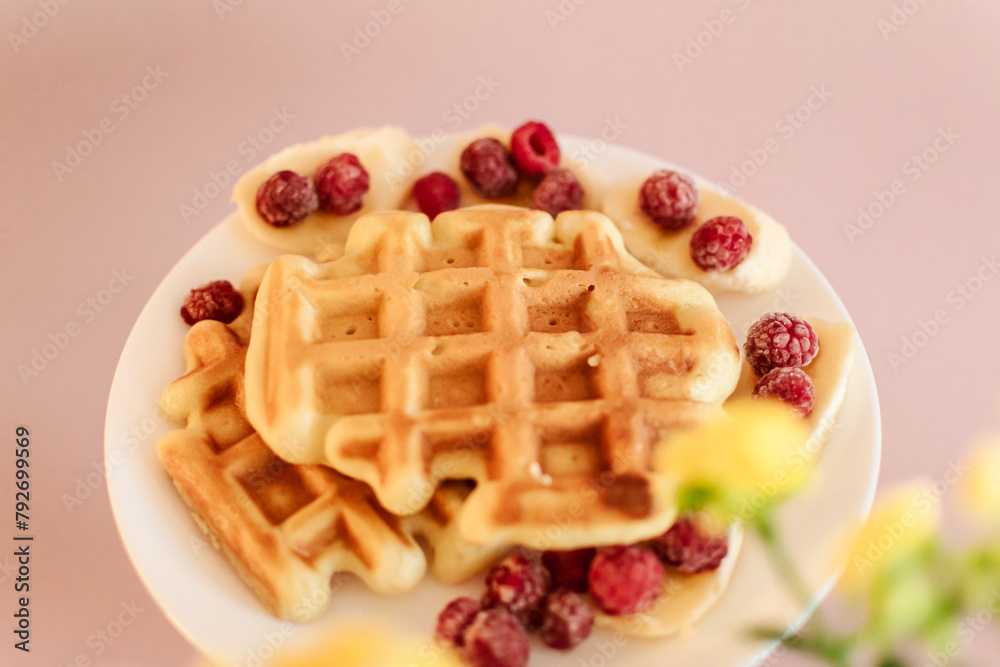 Viennese waffles with raspberries and bananas on a white plate next to yellow flowers in a pot. plain light background. homemade baking. recipe Viennese waffles with raspberries and bananas on a white plate next to yellow flowers in a pot. plain light background. homemade baking. recipe