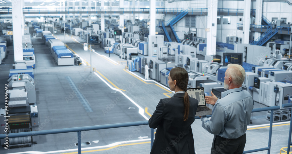 Middle Aged Engineer and Female Scientist Standing with Their Back, Using Laptop Computer and Talking in a Factory Facility with Equipment Producing Modern Technical Devices for Different Industries Middle Aged Engineer and Female Scientist Standing with Their Back, Using Laptop Computer and Talking in a Factory Facility with Equipment Producing Modern Technical Devices for Different Industries